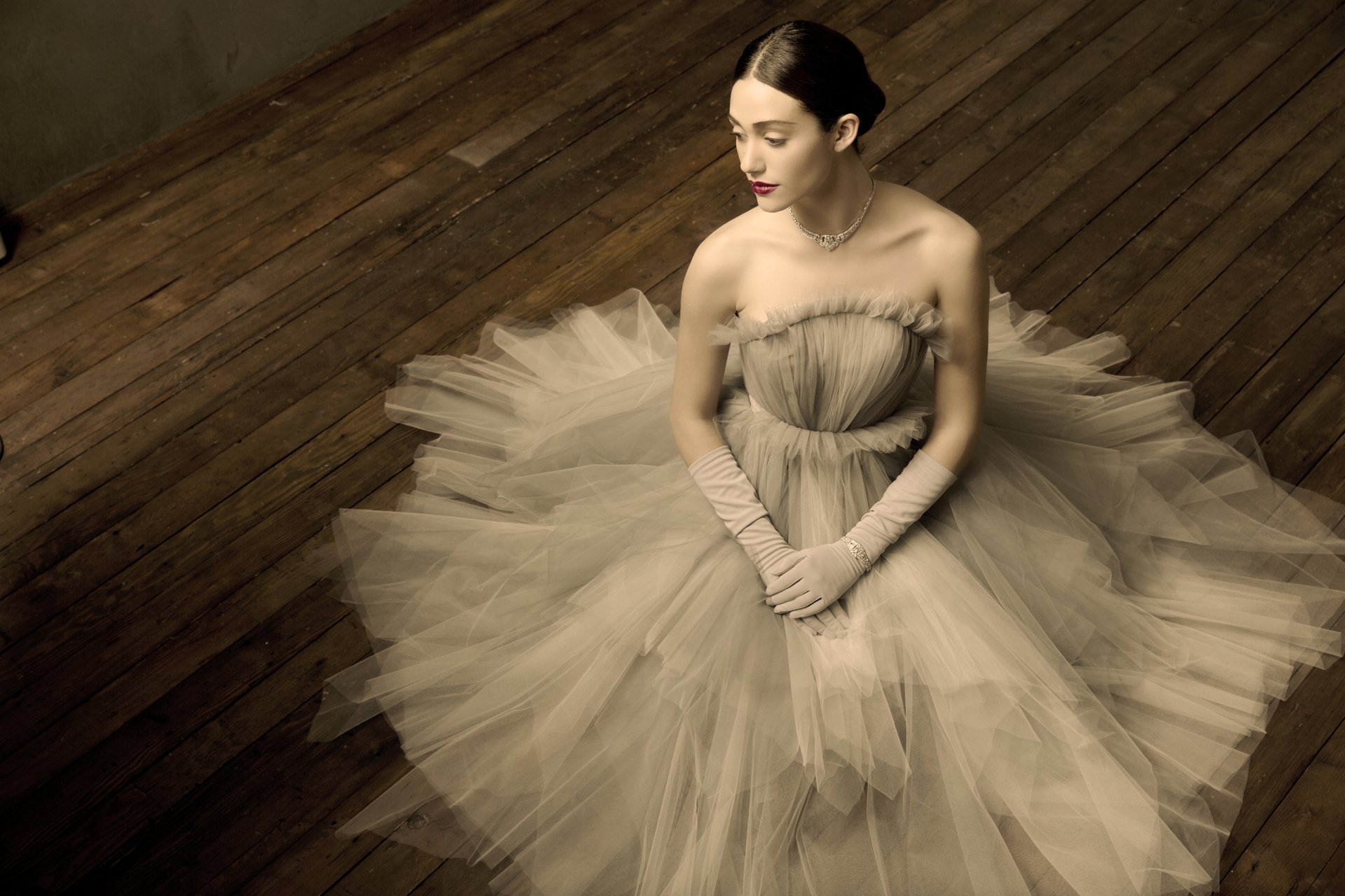 HD desktop wallpaper featuring celebrity Emmy Rossum in an elegant, flowing gown seated gracefully on a wooden floor.