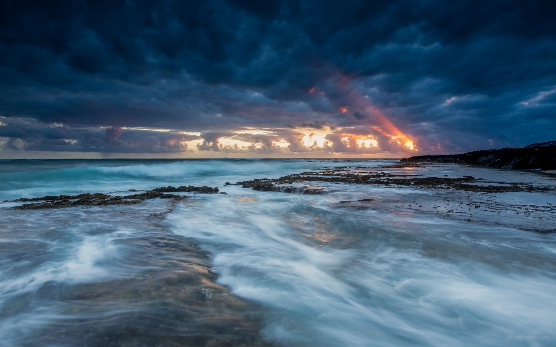HD desktop wallpaper featuring a dramatic coastline with dark clouds, waves crashing on rocky shores, and a vibrant sunset breaking through the stormy sky.
