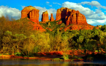 HD PC desktop wallpaper showing a vibrant canyon landscape with red rock formations, green trees, and a reflective water body under a blue sky with clouds.