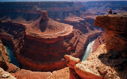 HD desktop wallpaper featuring the stunning Grand Canyon, showcasing its majestic cliffs and winding river. A person is sitting on a rock, enjoying the breathtaking natural landscape.