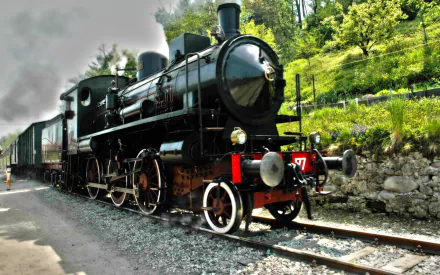 HD desktop wallpaper featuring a classic black steam train vehicle traveling on tracks beside a green hillside under a cloudy sky.