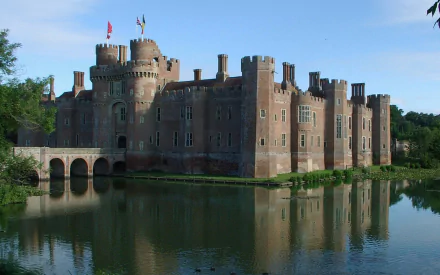 A high-definition PC desktop wallpaper featuring Herstmonceux Castle, a majestic man-made medieval fortress reflected in the calm surrounding water under a clear blue sky.