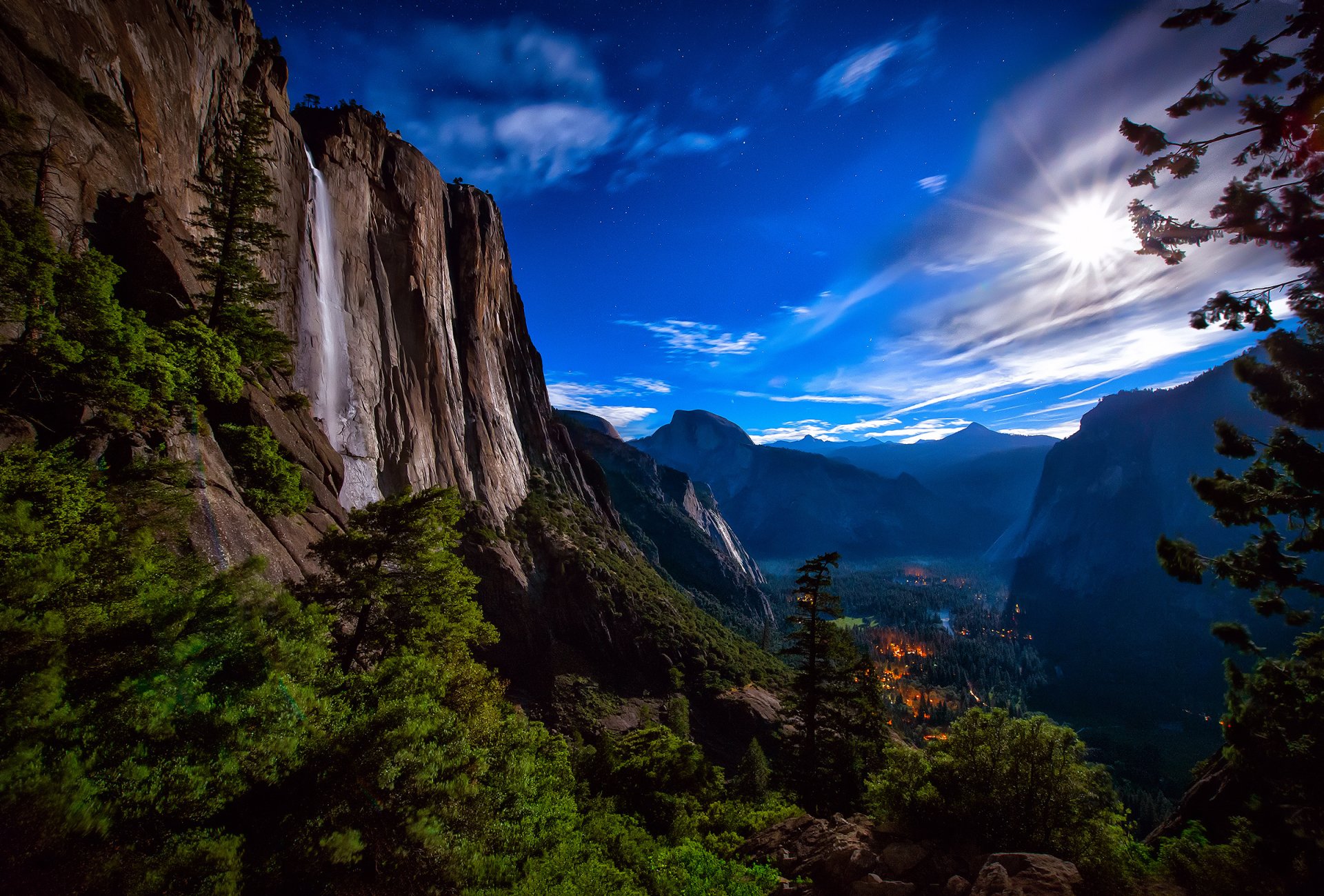 A stunning HD wallpaper capturing a majestic waterfall nestled among towering cliffs and lush greenery, under a vibrant blue sky and illuminated by sunlight.