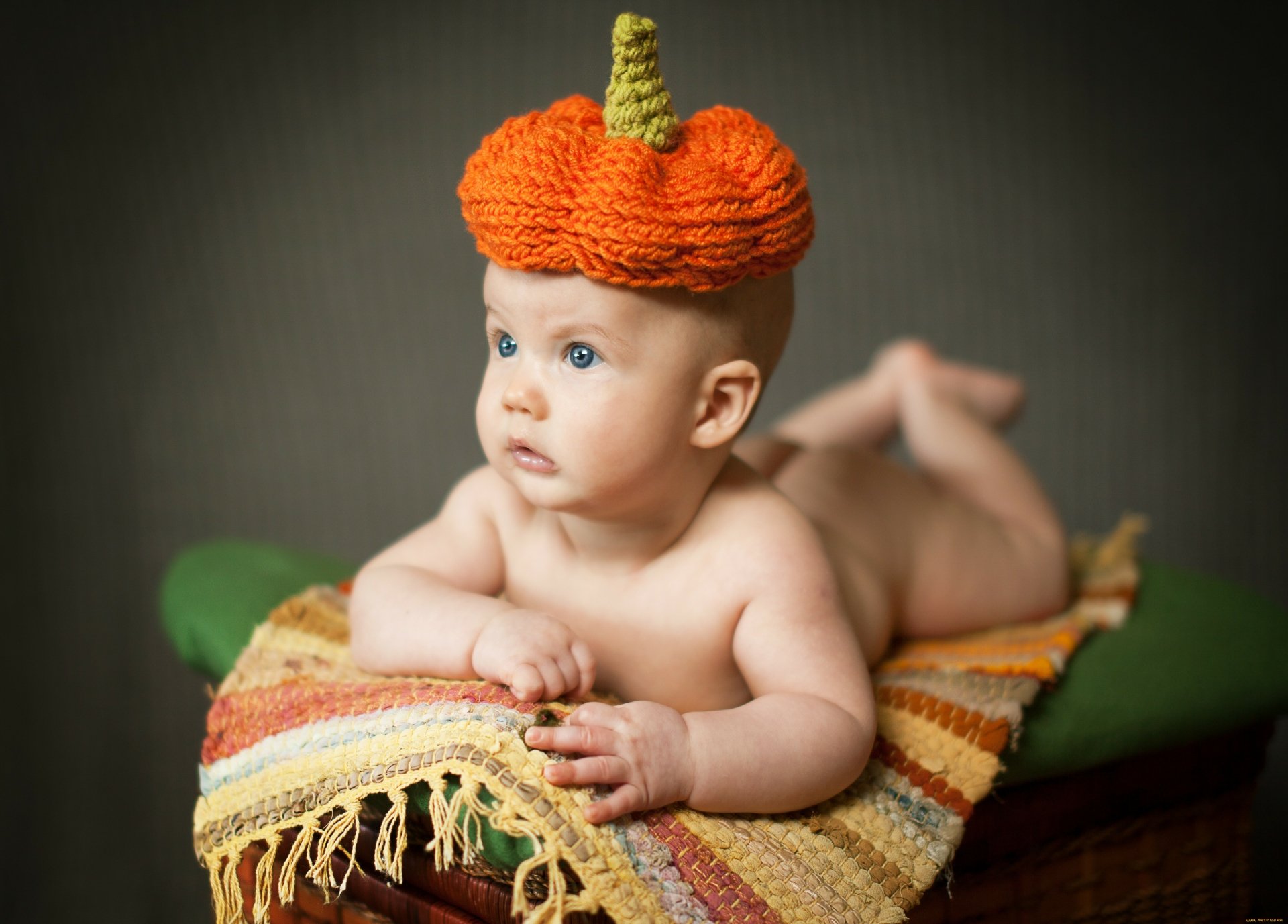 A 4K Ultra HD photo of a baby lying on a colorful woven blanket against a dark background, wearing a knitted orange pumpkin hat.