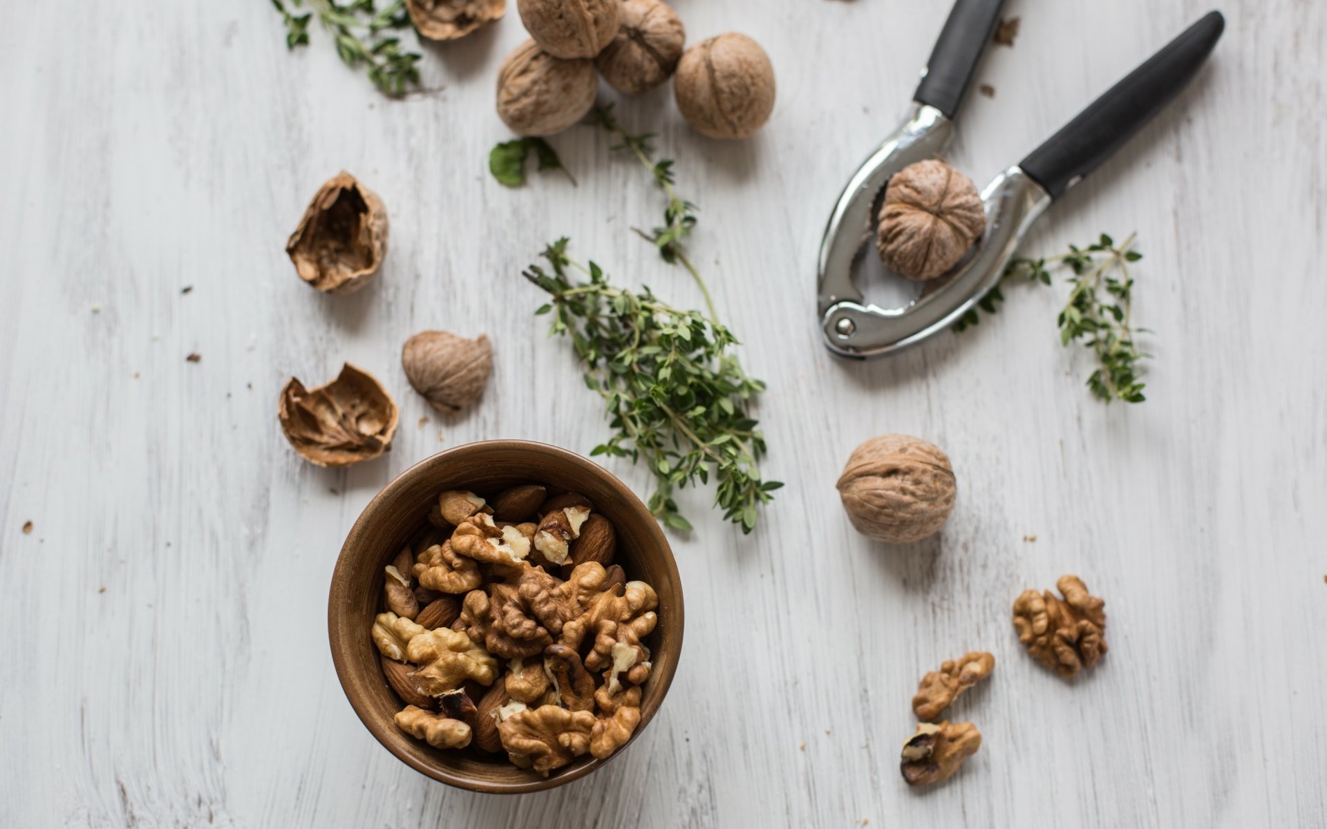 HD desktop wallpaper featuring a close-up of whole and cracked walnuts, a nutcracker, and fresh herbs on a light wooden surface.