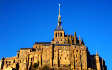 HD PC desktop wallpaper background of Mont Saint-Michel: the religious abbey rising from a sunlit rocky island against a vivid blue sky.