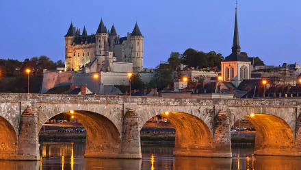 HD desktop wallpaper showcasing the illuminated Château de Saumur and a historic stone bridge reflecting in the calm river at dusk.