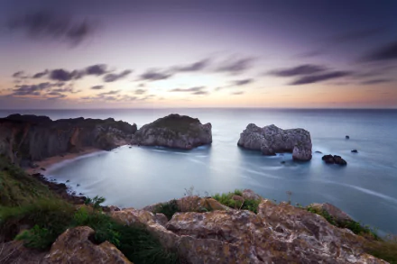 HD PC desktop wallpaper showing a serene coastline at dusk, with rocky cliffs, calm ocean waters, and a pastel-colored sky blending into the horizon.