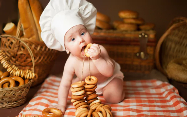 A baby wearing a chef’s hat sits on a checkered cloth, playfully holding and tasting a string of bagels, captured in vibrant 4K Ultra HD photography.