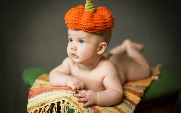 A 4K Ultra HD photo of a baby lying on a colorful woven blanket against a dark background, wearing a knitted orange pumpkin hat.