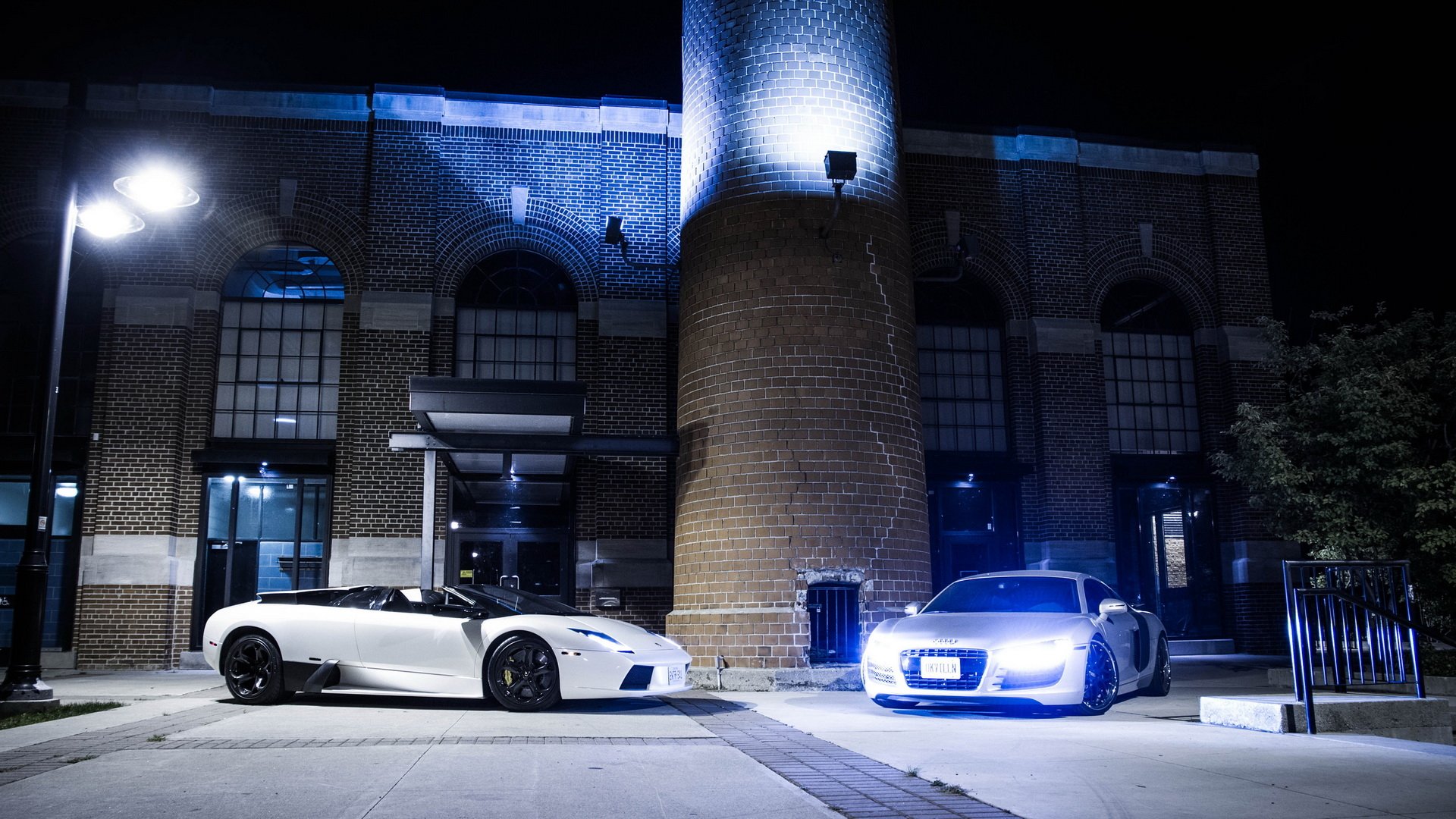 A sleek white Lamborghini and an Audi parked in front of a stylish brick building, illuminated by blue lighting at night, creating an eye-catching automotive scene.
