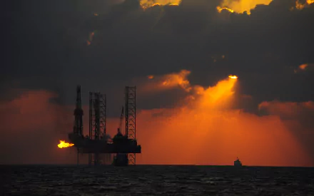 HD desktop wallpaper showing a man-made oil platform silhouetted against a dramatic sunset over the ocean with dark clouds and golden light beams.
