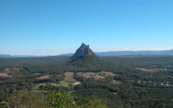 HD desktop wallpaper of the Glasshouse Mountains nature scene: a jagged volcanic peak rising from green farmland and eucalyptus forest beneath a clear blue sky.