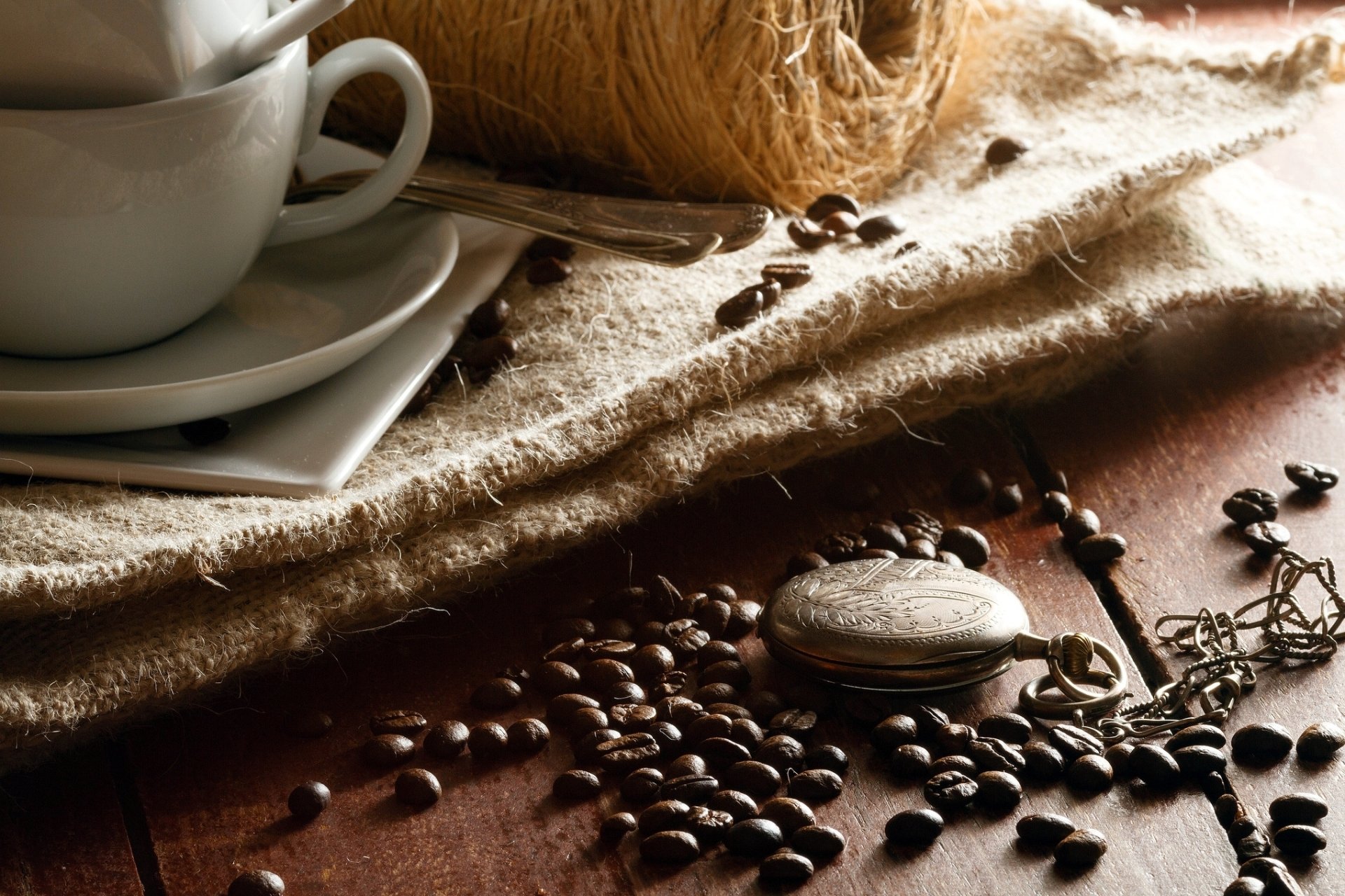 HD desktop wallpaper featuring a close-up of coffee beans scattered on a rustic wooden surface next to a white cup and saucer on a textured cloth.