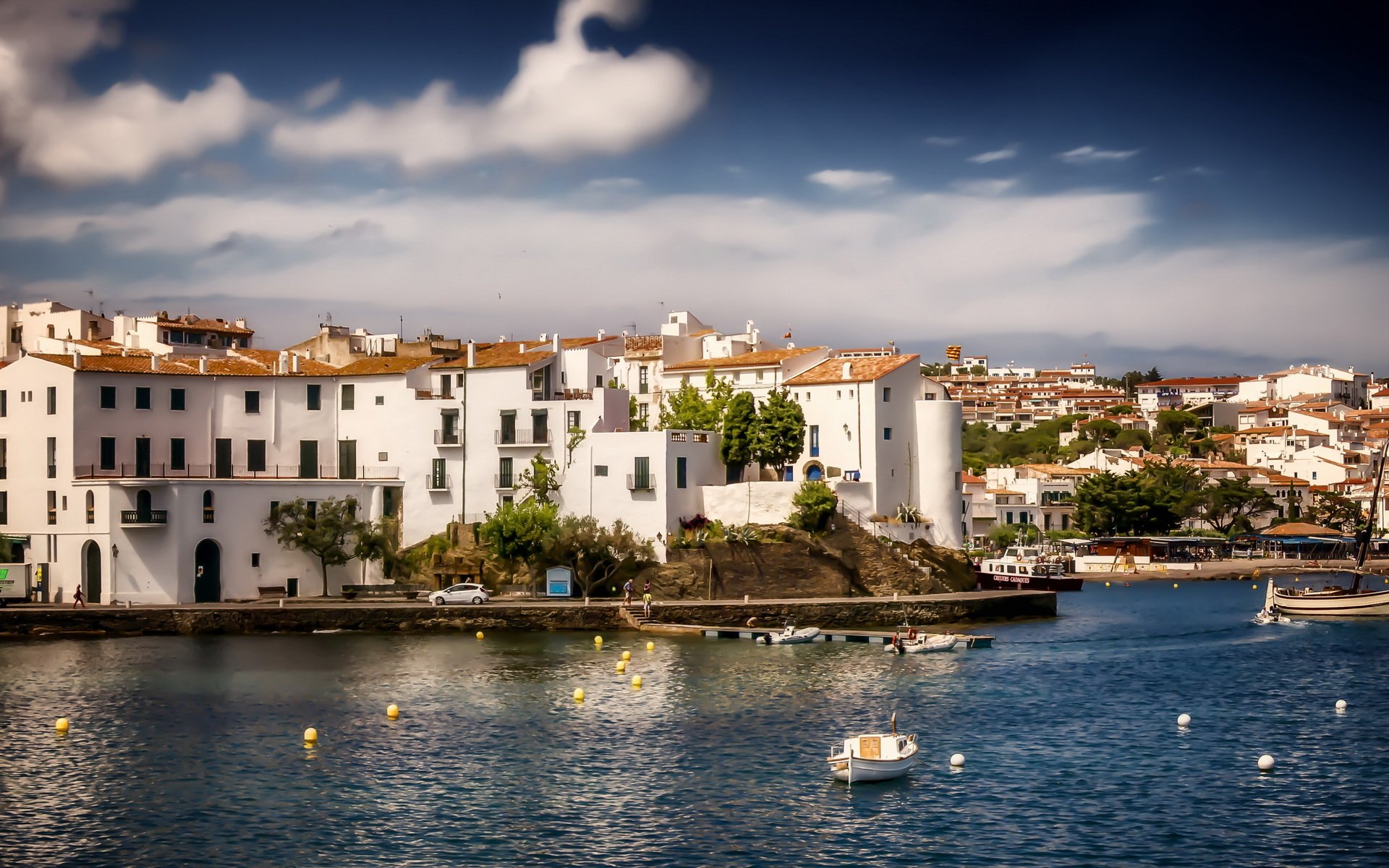 Man-made coastal buildings in Cadaqués bathed in sunlight, overlooking calm blue waters under a partly cloudy sky, captured in a high-definition PC desktop wallpaper.