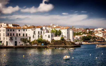 Man-made coastal buildings in Cadaqués bathed in sunlight, overlooking calm blue waters under a partly cloudy sky, captured in a high-definition PC desktop wallpaper.