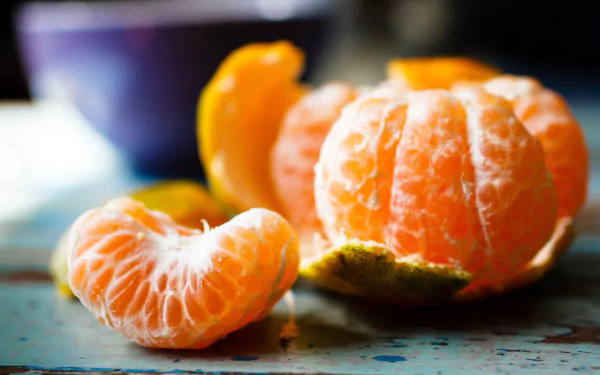 A vibrant HD wallpaper featuring a freshly peeled mandarin, showcasing its juicy segments and bright color, set against a rustic background with a bowl in the distance.