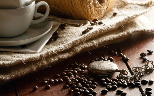 HD desktop wallpaper featuring a close-up of coffee beans scattered on a rustic wooden surface next to a white cup and saucer on a textured cloth.
