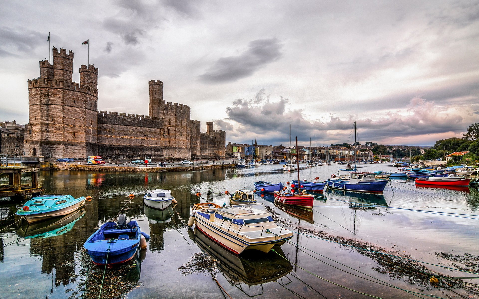 HD desktop wallpaper showing Caernarfon Castle beside a harbor filled with colorful boats under a cloudy sky, highlighting this man-made historic site.