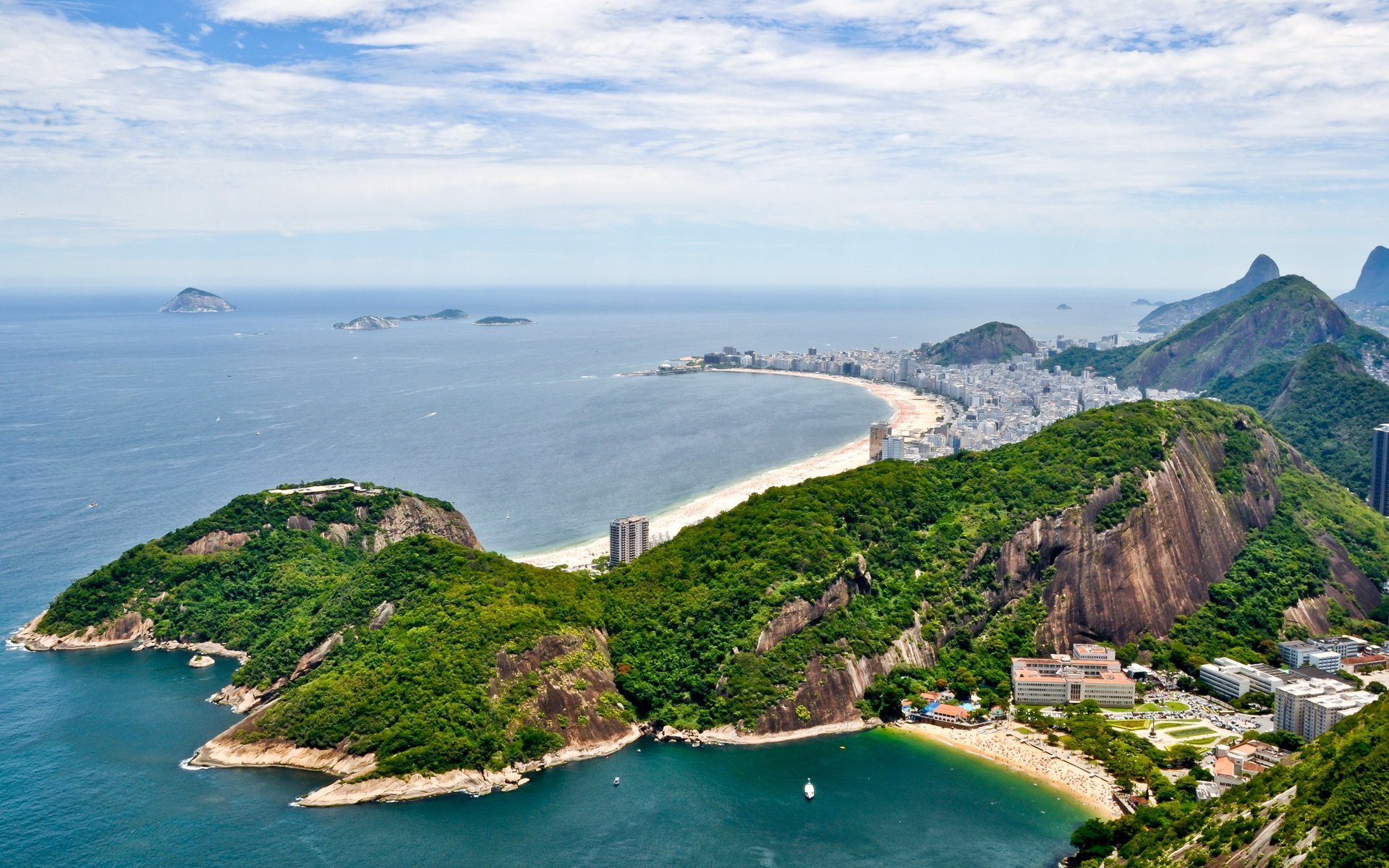 A HD desktop wallpaper showing the man-made cityscape and natural coastline of Rio de Janeiro under a partly cloudy sky.