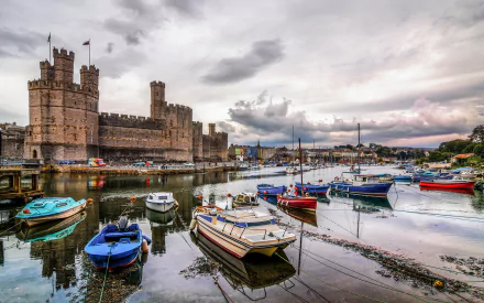 HD desktop wallpaper showing Caernarfon Castle beside a harbor filled with colorful boats under a cloudy sky, highlighting this man-made historic site.