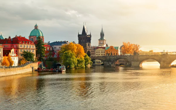 HD desktop wallpaper featuring the man-made Charles Bridge over a calm river, with historic buildings and autumn trees illuminated by soft, golden sunlight.