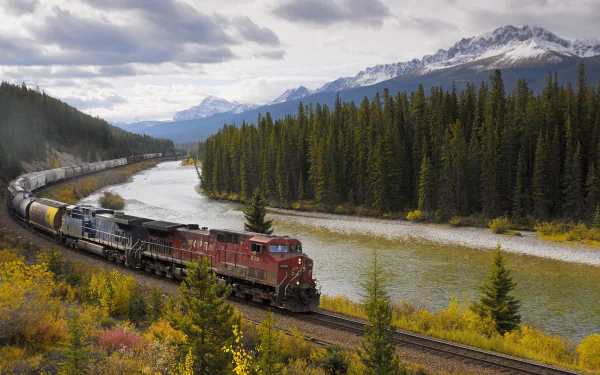 HD wallpaper of a red train traveling along a river with densely forested banks and snow-capped mountains in the background.