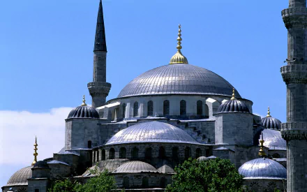 A stunning HD desktop wallpaper of the Sultan Ahmed Mosque, showcasing its iconic domes and minarets against a clear blue sky, surrounded by lush greenery.