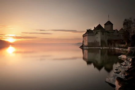 A serene sunset casts a warm glow over the calm waters near Château de Chillon in Veytaux, Switzerland, reflecting the castle and the tranquil scenery.