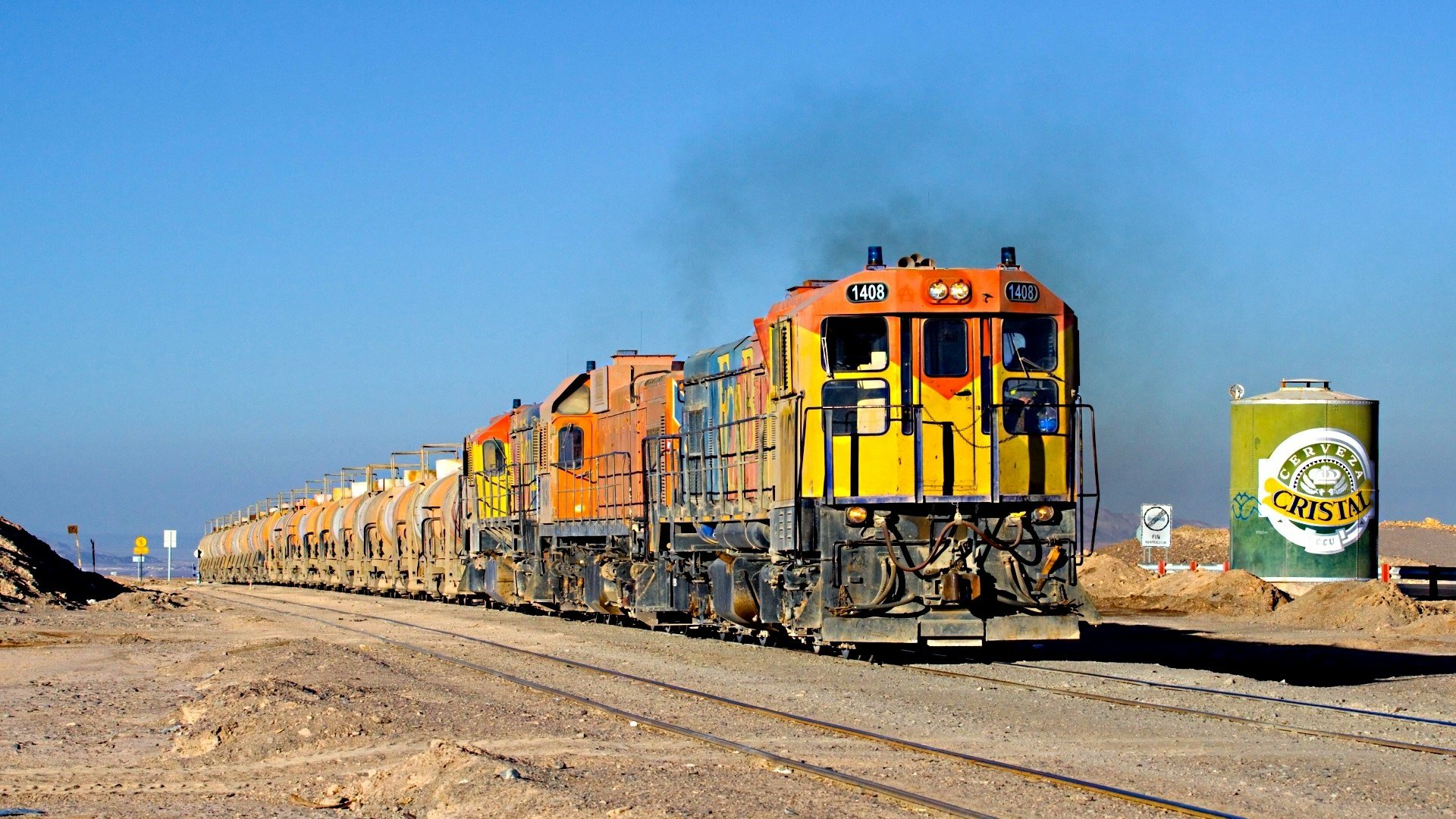 HD desktop wallpaper featuring a vibrant orange and yellow freight train traveling on tracks through a desert landscape under a clear blue sky.