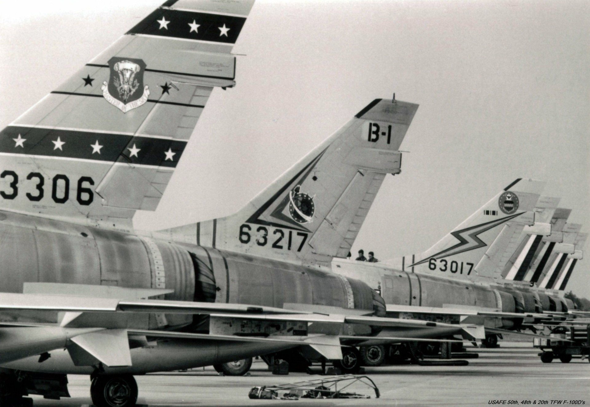 Military 2K Quad HD desktop wallpaper of lined-up North American F-100 Super Sabre tail sections in black-and-white, showing unit markings and serial numbers on an airfield tarmac.