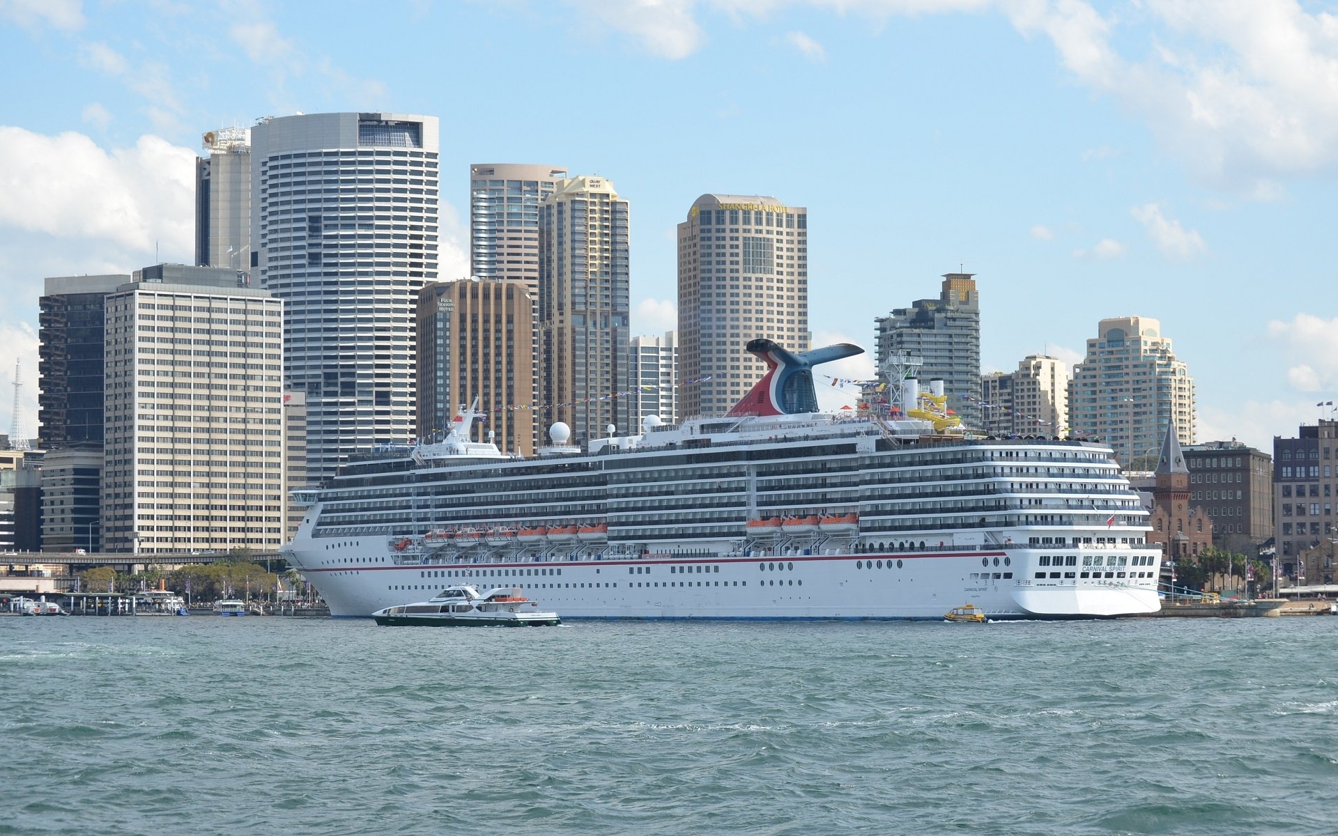 A cruise ship, Carnival Spirit, sails in Sydney Harbour, with the city's skyline towering in the background, showcasing vibrant buildings and a stunning Australian waterfront.
