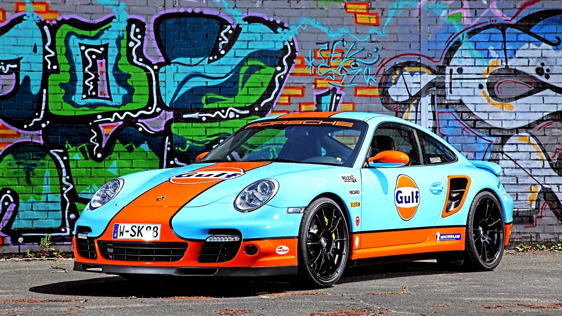 HD PC desktop wallpaper featuring a Porsche sports car in Gulf racing livery parked against a vibrant graffiti-covered brick wall.