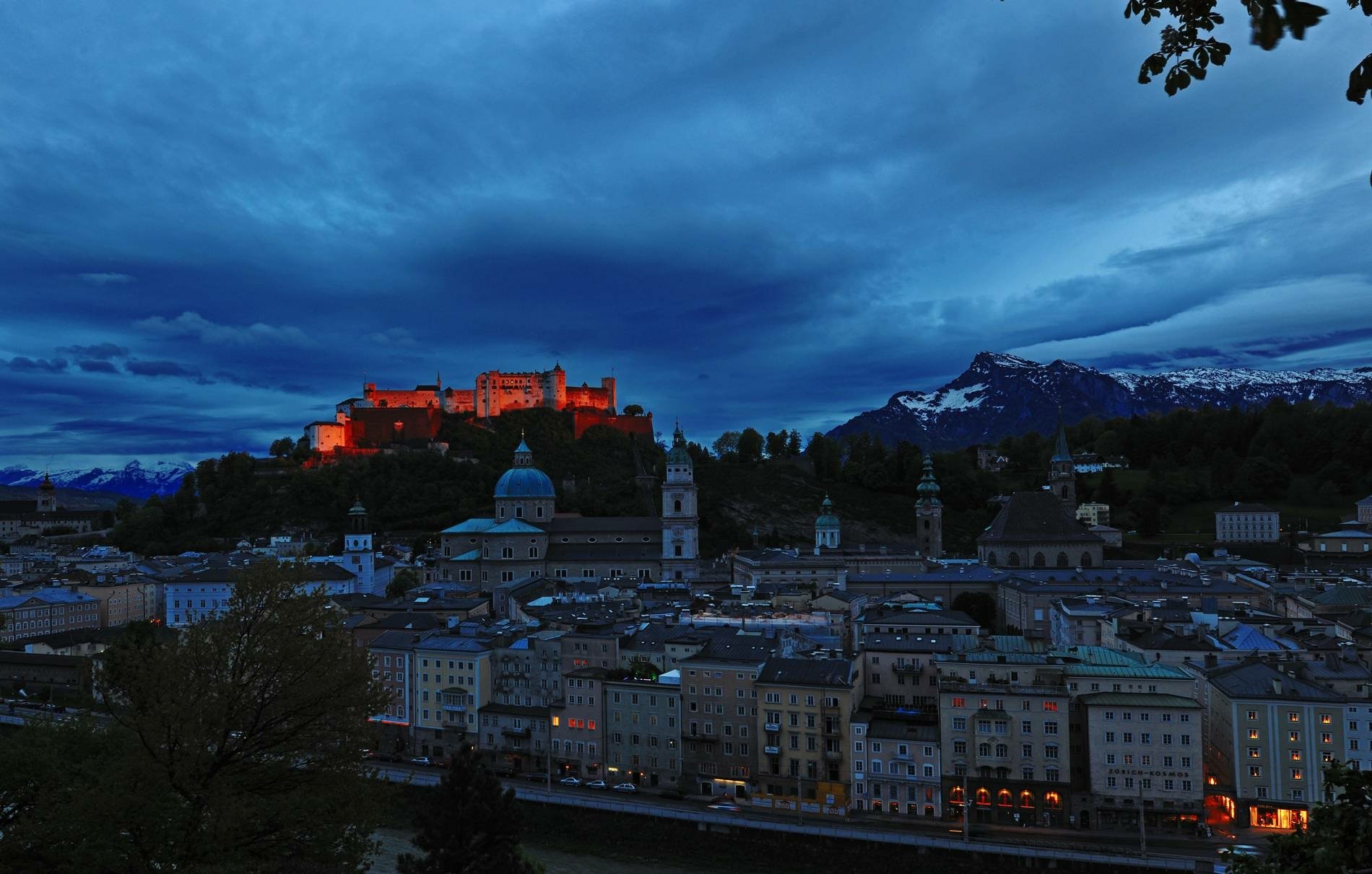 HD desktop wallpaper: old man-made Hohensalzburg Castle glowing above the dark night city, warm lights contrasting the deep blue sky and distant snowy peaks.