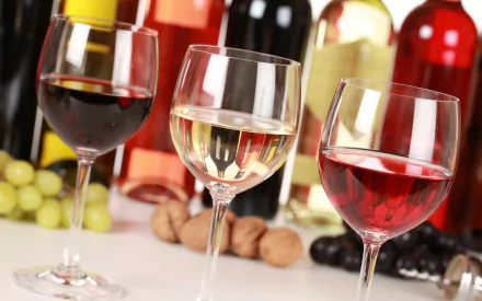 A vibrant HD desktop wallpaper featuring three wine glasses—red, white, and rosé—set against a colorful backdrop of wine bottles and fresh grapes.