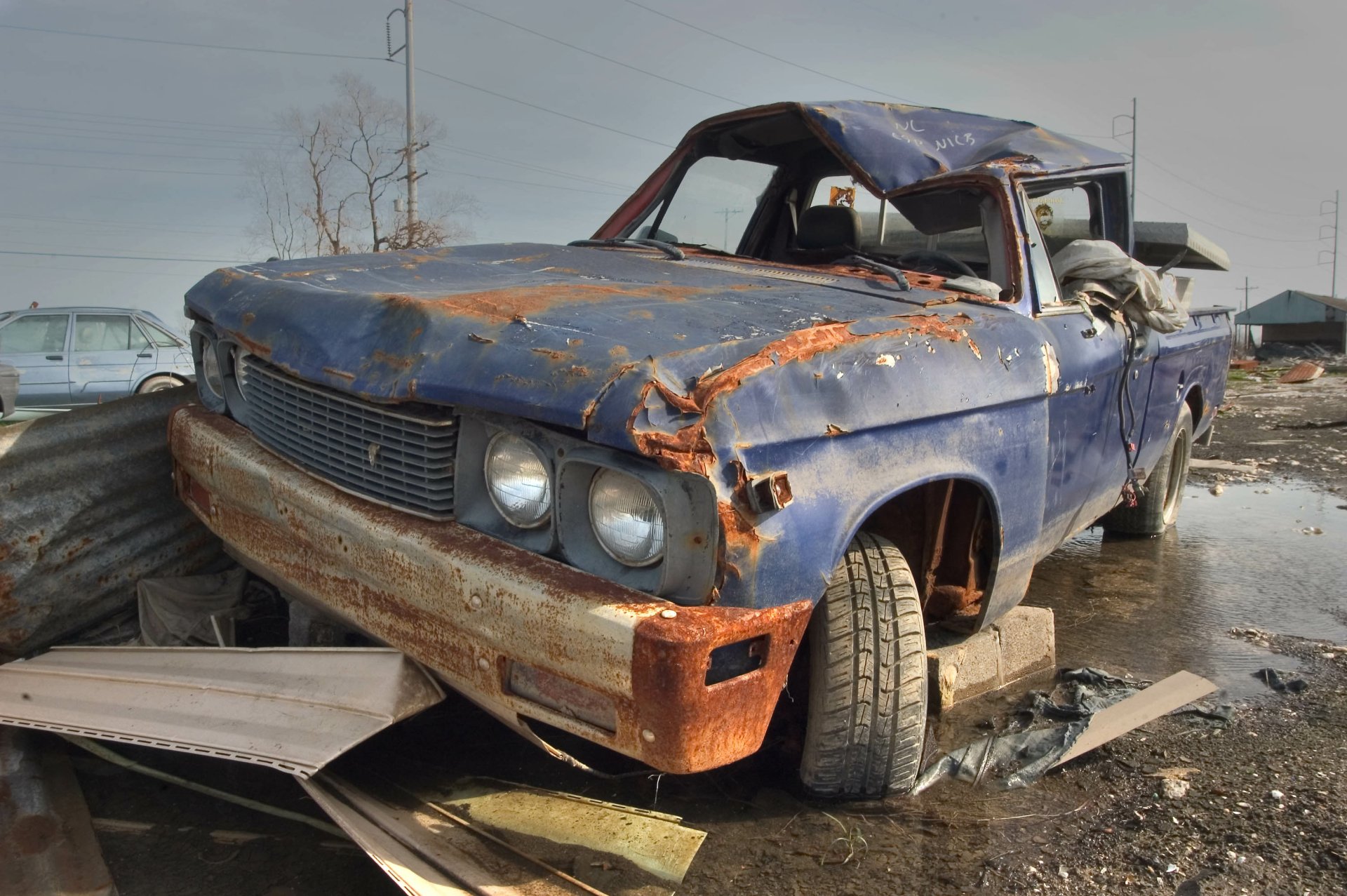 2K Quad HD PC desktop wallpaper showing an old wrecked blue vehicle in a muddy junkyard, rusted panels and a flat tire.