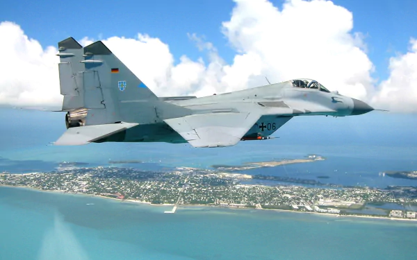 A Mikoyan MiG-29 jet fighter soars above a coastal landscape, showcasing its sleek design against a backdrop of vibrant blue skies and tranquil waters.