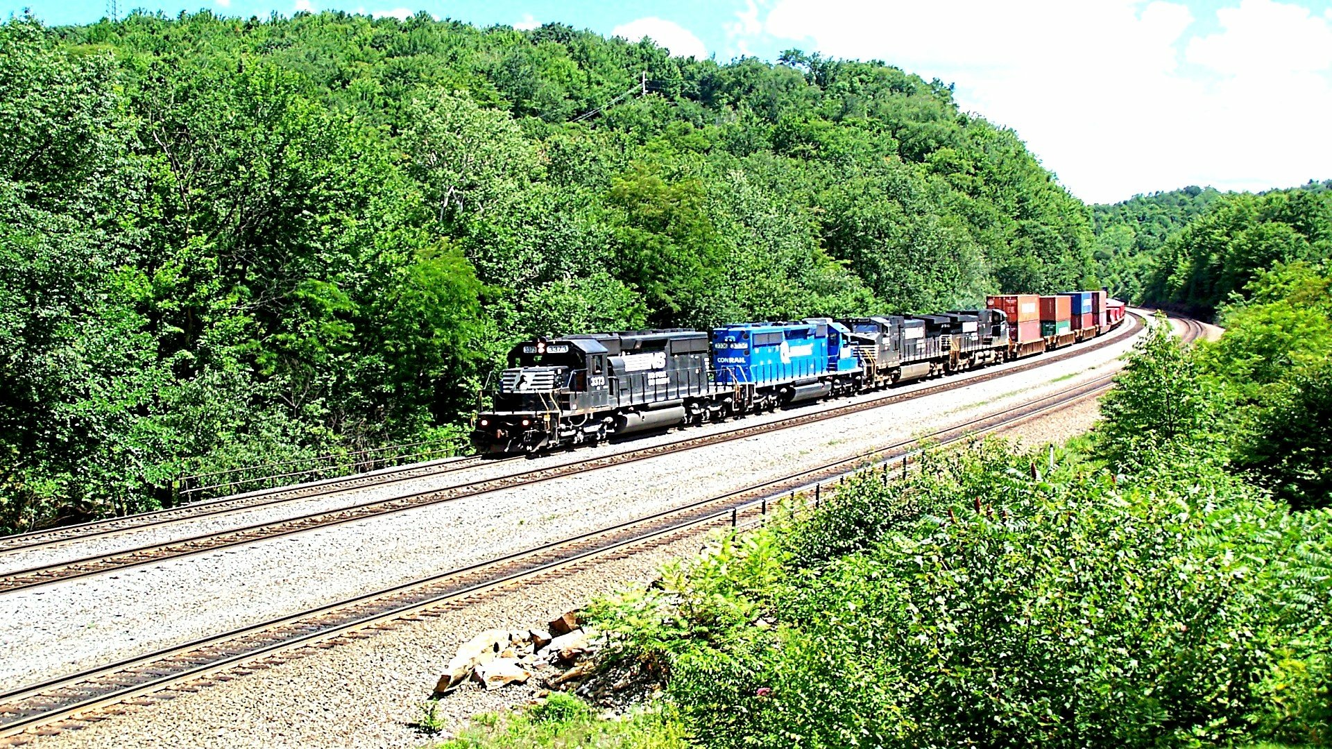 HD PC desktop wallpaper and background: a freight train vehicle in black and blue curves through a lush green forest along parallel tracks under a bright sky.