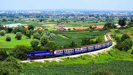 A vibrant countryside landscape features a blue train winding through lush fields, framed by distant hills and a charming village, creating an inviting HD wallpaper.