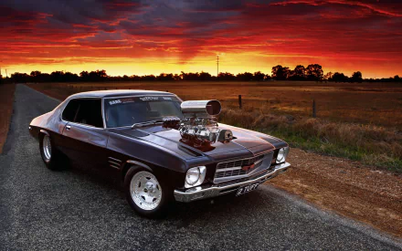 HD desktop wallpaper featuring a classic Holden Monaro muscle vehicle parked on a rural road at sunset with a vibrant red sky background.