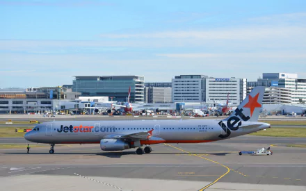A Jetstar Airbus A321 taxiing on the runway at an airport, with modern terminal buildings in the background, captured in a vibrant HD image.