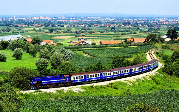 A vibrant countryside landscape features a blue train winding through lush fields, framed by distant hills and a charming village, creating an inviting HD wallpaper.