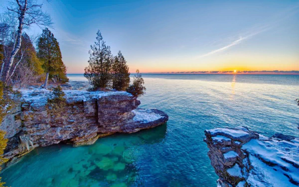 HD desktop wallpaper: Lake Michigan sunrise over a rocky coastline with evergreen trees, clear turquoise water and a glowing horizon.