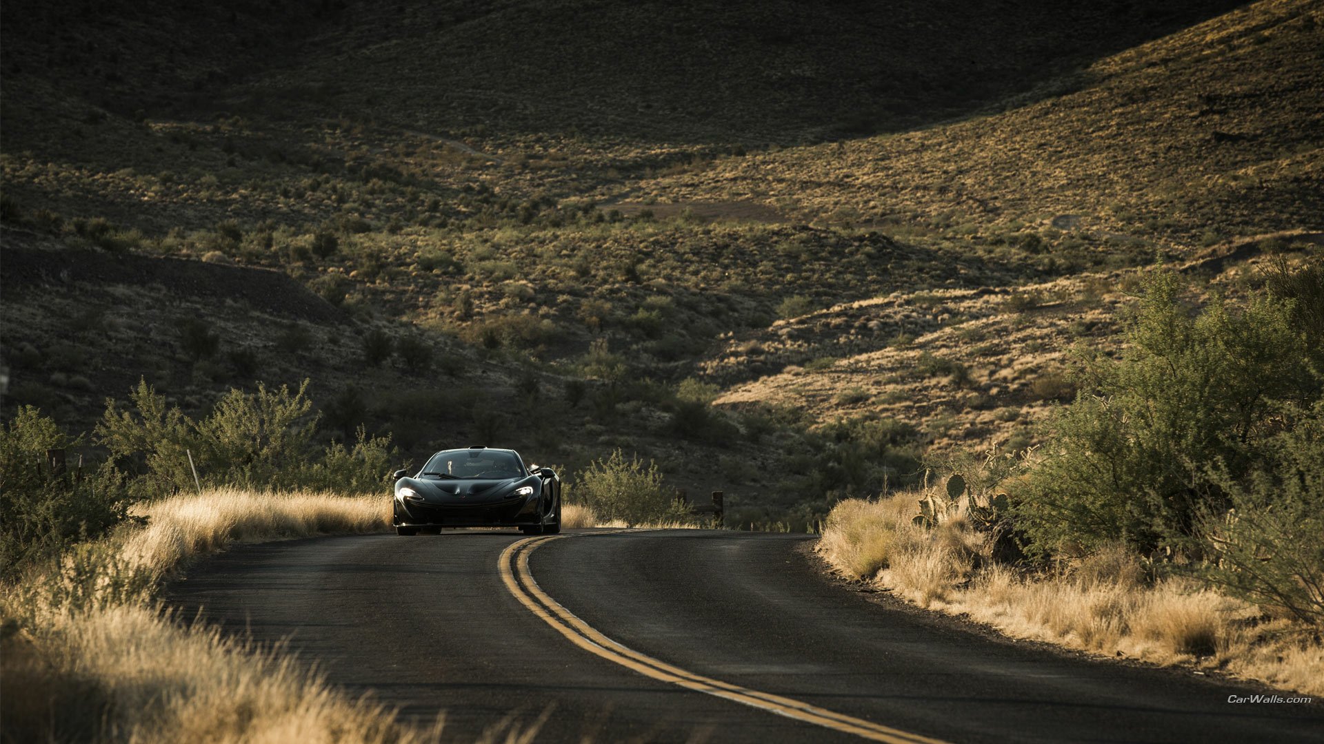HD PC desktop wallpaper featuring a McLaren P1 driving on a winding road through a sunlit, rugged landscape.
