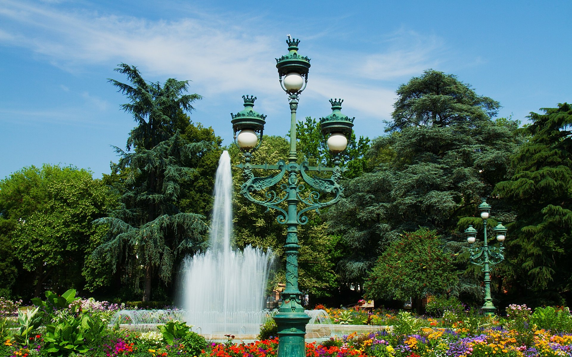 HD desktop wallpaper showcasing a vibrant park scene with ornate lampposts, a flowing water fountain, colorful flowerbeds, and lush green trees under a clear blue sky.