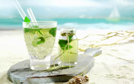 HD desktop wallpaper featuring a refreshing cocktail with mint and ice by the beach, accompanied by a small jar with a green drink.