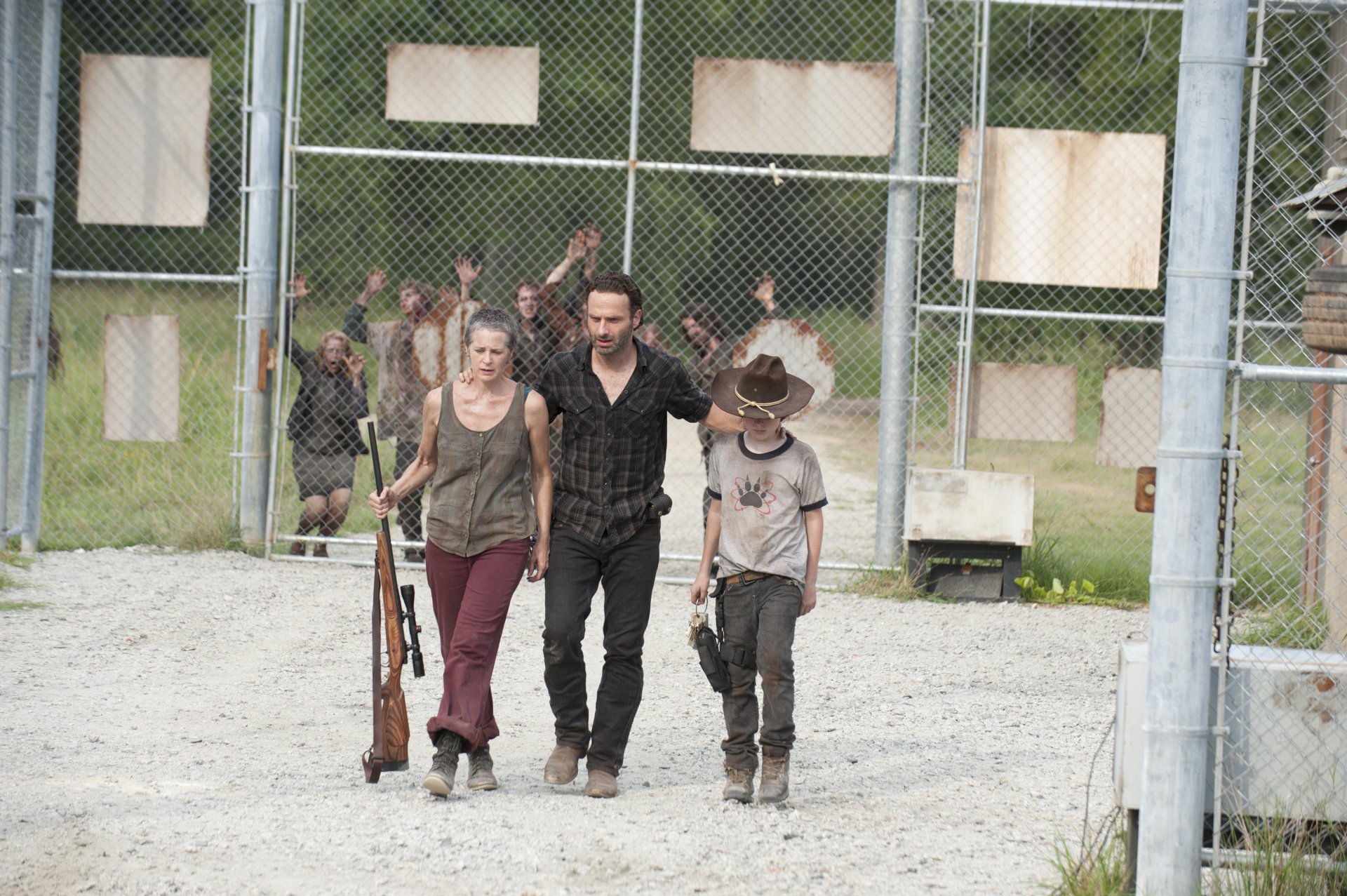 Rick Grimes, Carl Grimes, and Carol Peletier walk confidently through a fenced area, with walkers in the background, from the TV show The Walking Dead. HD wallpaper features main characters.