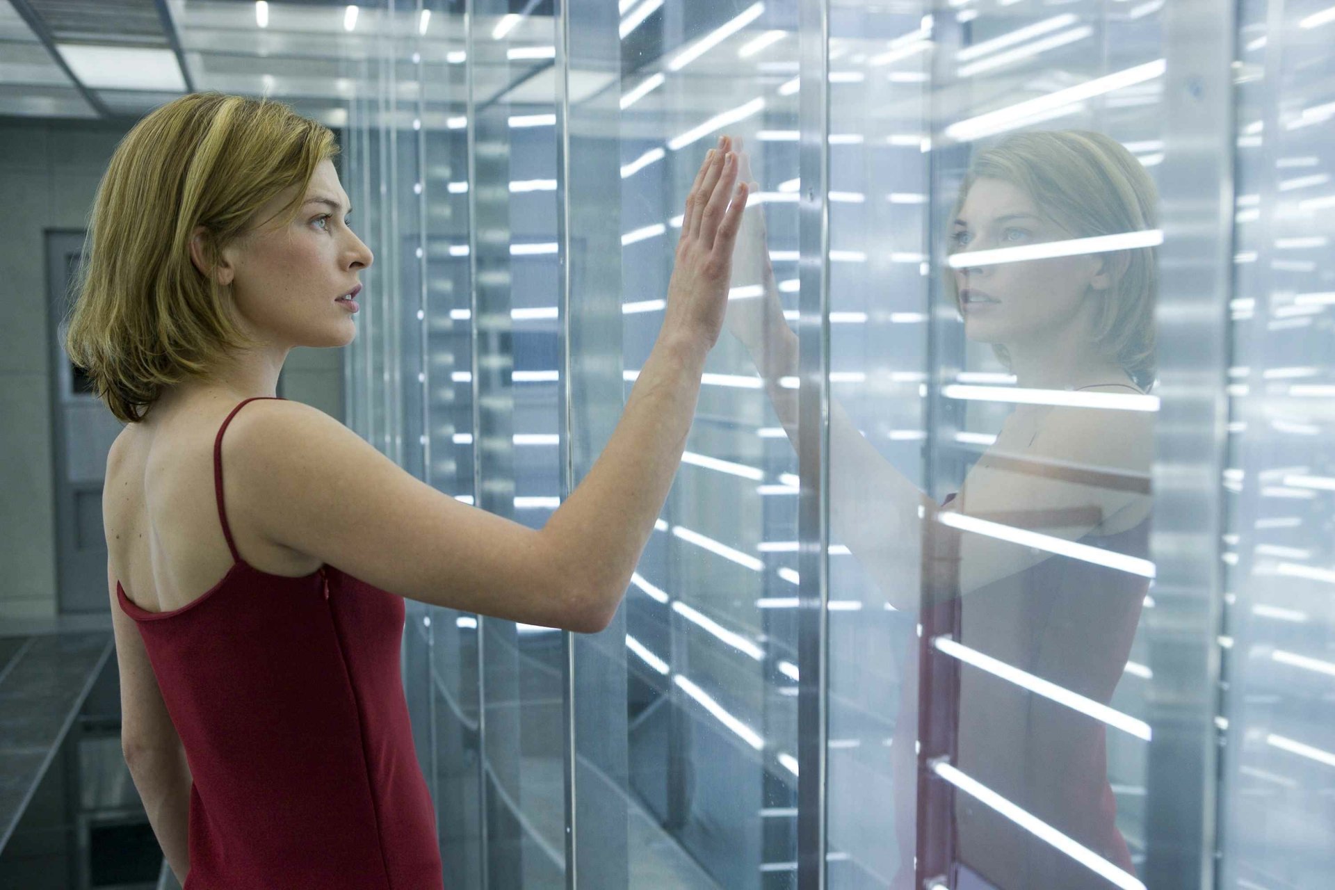 Milla Jovovich stars in this captivating scene from the movie Resident Evil, showcasing her character reaching out to a reflective surface in a high-tech environment. HD desktop wallpaper.