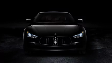 A sleek Maserati Ghibli in black, illuminated by its headlights, stands out against a dark background, showcasing the vehicle's elegant design in this HD desktop wallpaper.