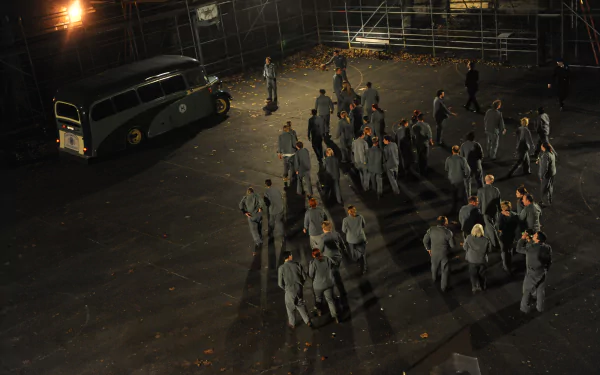 A scene from *The Shawshank Redemption*, featuring a group of men in gray suits gathered on a dimly lit prison yard, with buses seen in the background. High-definition wallpaper.